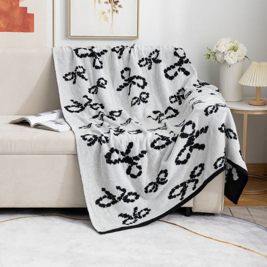 A gray and black knit throw blanket with bow patterns, draped over the arm of a beige sofa.