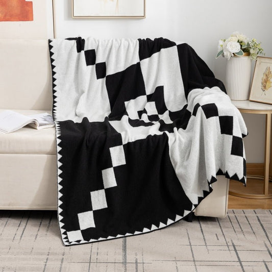 A black and white geometric patterned fleece knit blanket draped over the arm of a sofa, with a part of the blanket folded back to reveal a cream-colored surface underneath.