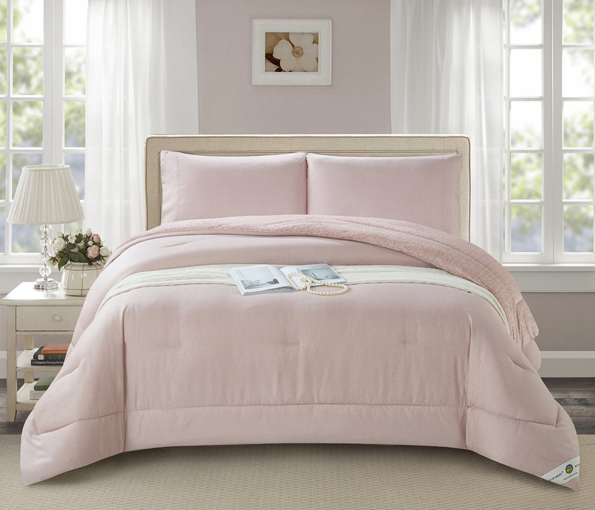 A blush pink minimalist quilted comforter set on a bed in a room with a window and lamp in the background.