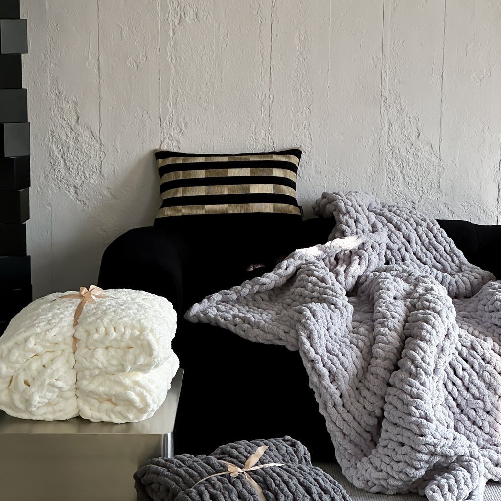 A cozy chunky knit blanket in gray and white colors is draped over a armchair, with a cushion beside it. A rolled-up blanket with a bow on it is also visible in the foreground.