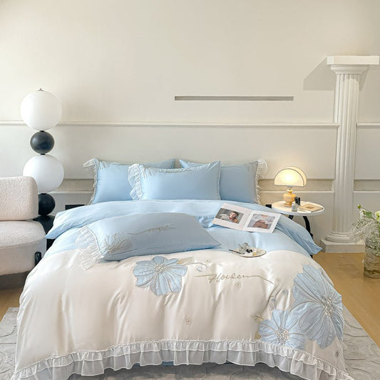 A bedding set with blue floral embroidery on a white background displayed on a bed, with a book, lamp, and decorative room elements visible in the background.