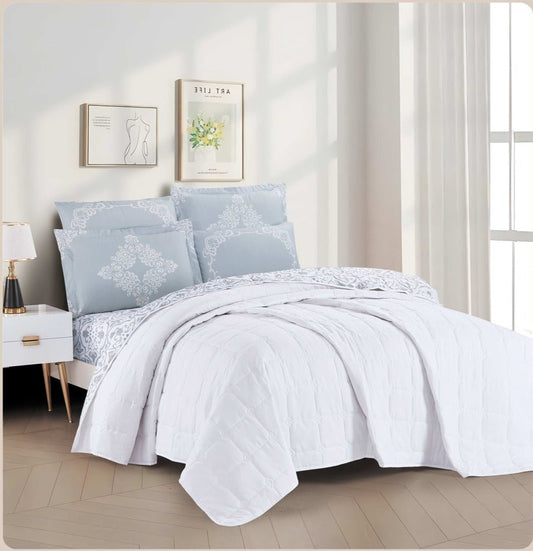 A white quilted bedding set with a botanical print, displayed on a bed in a room with decorative pillows, bed sheets, and a window.