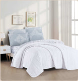A white quilted bedding set with a botanical print, displayed on a bed in a room with decorative pillows, bed sheets, and a window.
