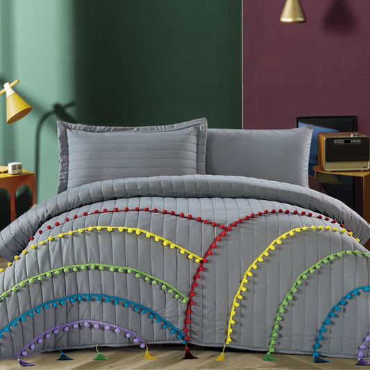 A gray comforter with multicolored pom pom fringe spread across the edges, displayed in a bedroom setting with decorative pillows, bed sheets, and a quilt.