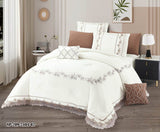 A white comforter set with floral lace detailing, displayed in a bedroom setting with brown accent pillows and a matching white and brown valance.