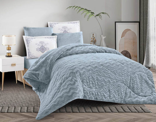A quilted wave-pattern winter flannel comforter set in blue, displayed on a bed with matching pillows and bed sheet, in a room with wooden flooring and a plant.