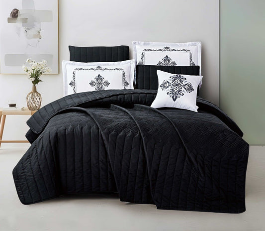 A black striped quilted bedspread on a bed, with decorative pillows on top, in a room with a light grey wall and a wooden floor.
