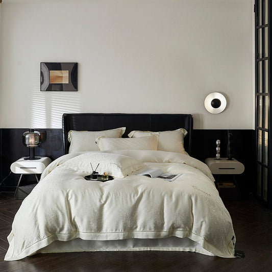 An ivory white duvet cover set on a bed, with decorative pillows, a book, and a remote control visible on the bed. The room has a minimalist design with a framed picture on the wall and a bedside lamp on the tables.