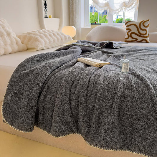 A gray sherpa knit throw on a bed with a book and a water bottle on top, in a room with a window and a cushion in the background.