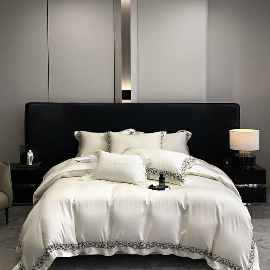 A white duvet cover set with embroidered detailing, displayed on a bed with a black headboard, in a room with a gray wall color and a lamp on the side table.