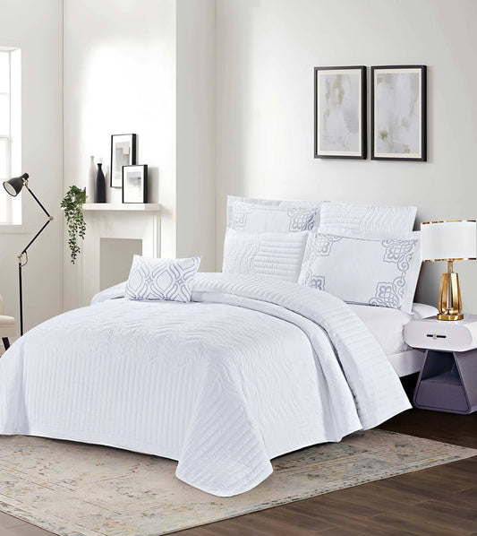 A white, quilted bed cover displayed on a bed in a bright room, accompanied by decorative pillows and a reading lamp.