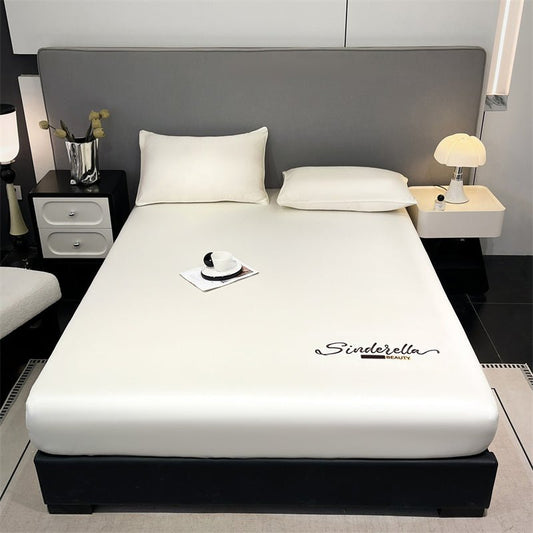 A white, embroidered fitted sheet on a bed with pillows, in a room with a nightstand and a lamp.