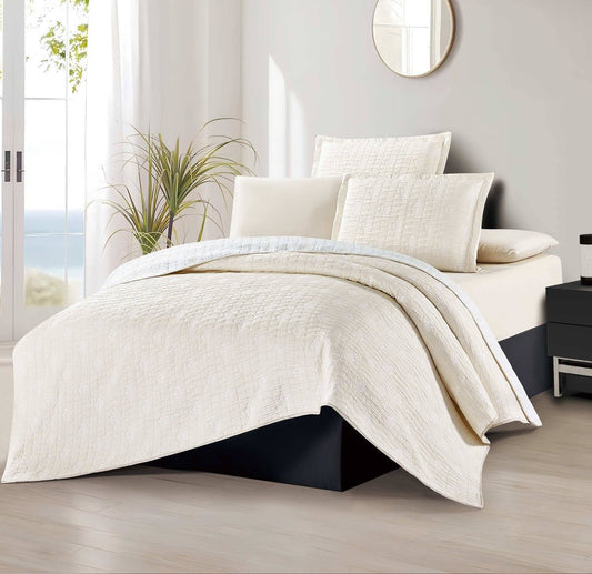 A beige bedspread displayed on a bed in a room with a plant and a mirror in the background.