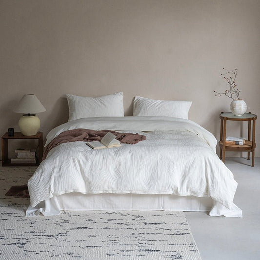 A white bedding set with stripes on a bed in a tidy bedroom, accompanied by a nightstand with a lamp and a decorative object, and a chair with a decorative item on top.