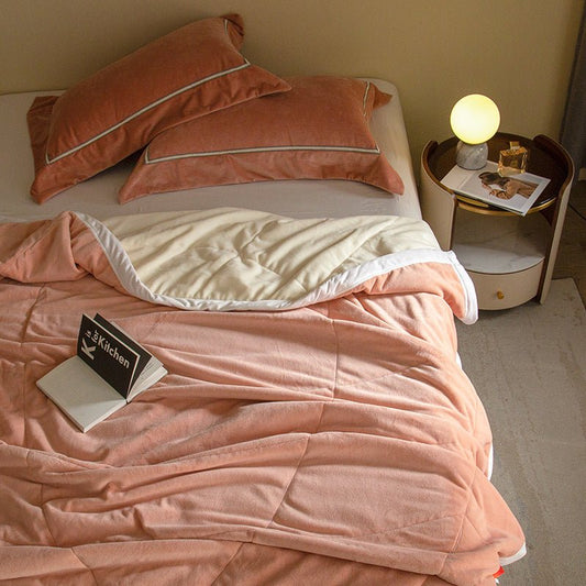 A coral fleece blanket draped over a bed with a book on top and a lit lamp beside the bed.