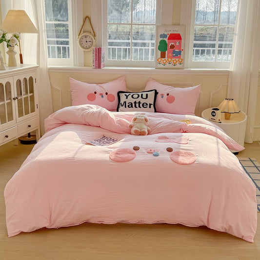 A pink bedding set with pig appliqués on a bed in a room with a window, clock, and decorative items.
