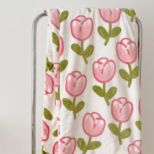 A blanket with a white background featuring a pattern of pink watercolor tulips and green leaves, draped over a metal rack.