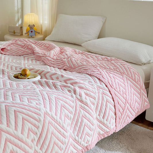 A pink and white geometric patterned blanket on a bed in a bedroom setting, with a bowl and apple on the bed, and a teddy bear and lamp in the background.