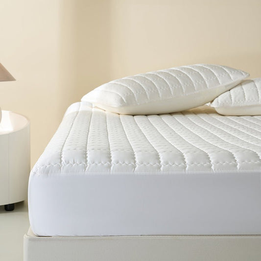 A white, breathable cloud sheet fitted on a bed with a pillow on top, in a room setting with a lamp and nightstand visible.