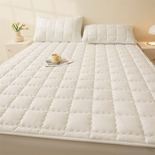 A white, quilted mattress pad on a bed with two pillows, a cup, and a floral arrangement in the background.