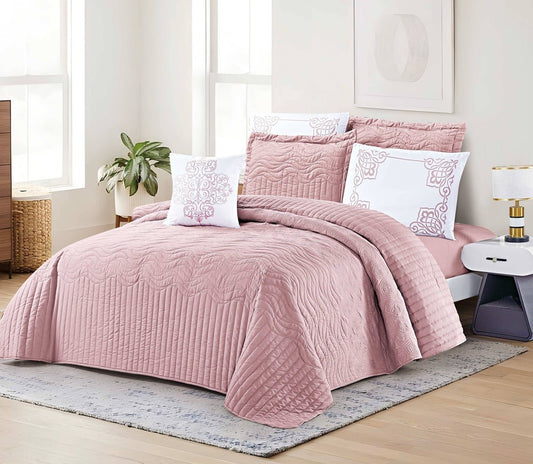 A pink quilt with a 3D ripple design displayed on a bed, accompanied by matching pillows and placed in a room with a light interior.