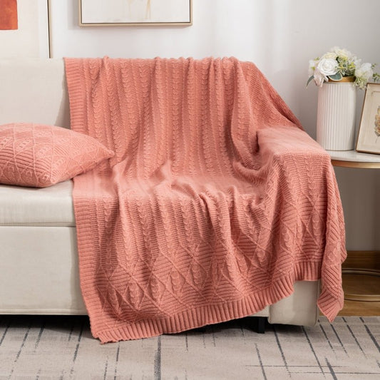 A pink HeritageKnit vintage cable blanket draped over the back of a sofa, with a matching pink pillow beside it, in a room with a wooden floor and a white wall in the background.