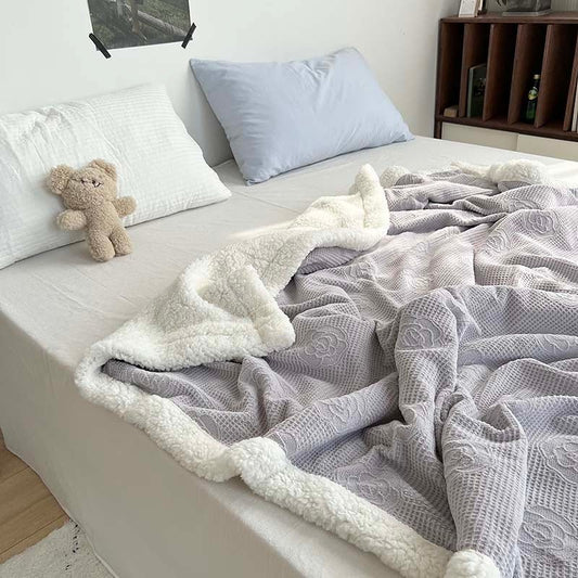 A floral knit blanket in gray and white, draped over the edge of a bed, with a teddy bear and pillows in the background.