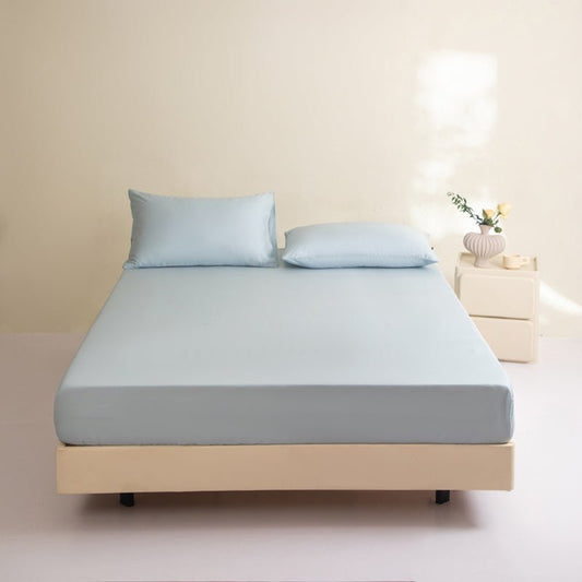 A blue fitted sheet on a bed, with two pillows on top of the sheet. The bed is placed on a wooden floor against a wall, with a small table and a plant to the side.