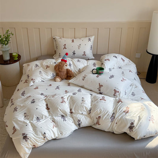 A white duvet cover set with a printed design featuring plushie figures, displayed on a bed with a teddy bear and a cup.