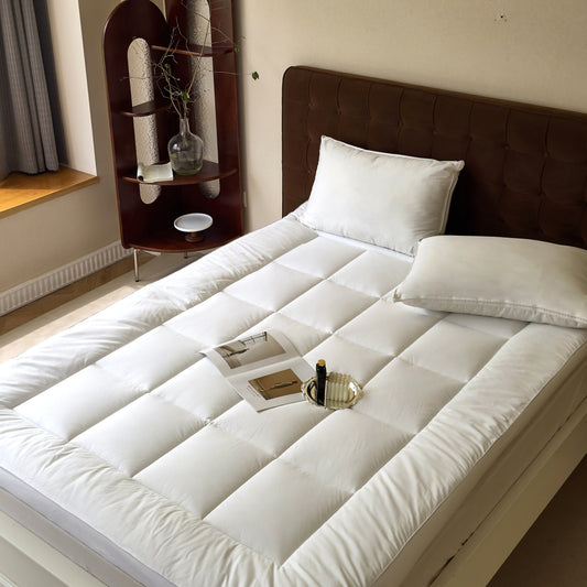 A white organic cotton mattress pad on a bed, with a pillow and a magazine on top, in a room with a wooden headboard and a nightstand.