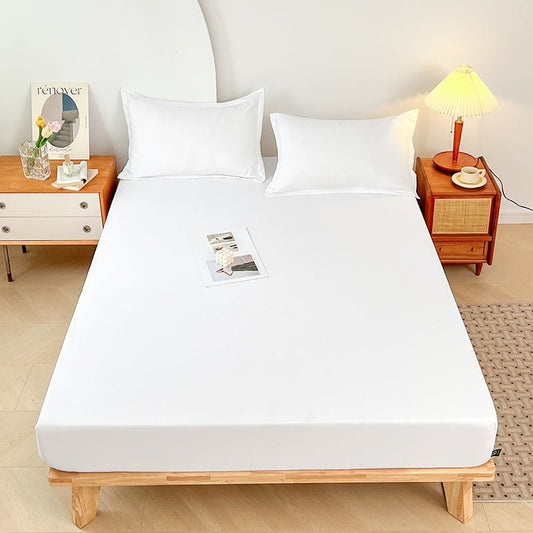 A white, triple-layer waterproof fitted sheet on a bed, with pillows and a book on the bed, placed in a room with a wooden bed frame, a nightstand, and a lamp.