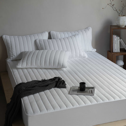 A white, waterproof, quilted fitted sheet on a bed, with several pillows on top and a black blanket draped over the side. A cup and a book are also visible on the bed.