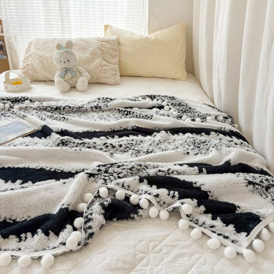 A black and white 3D textured throw with pom-pom details, laid out on a bed with pillows and a teddy bear in the background.