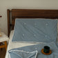 A blue silk pillow on a bed with a wooden headboard, the pillow has a solid pattern with what appears to be a natural fiber border.
