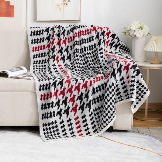 A houndstooth patterned throw blanket in black, white, and red, draped over the arm of a sofa in a room with a white carpet and various home decor.