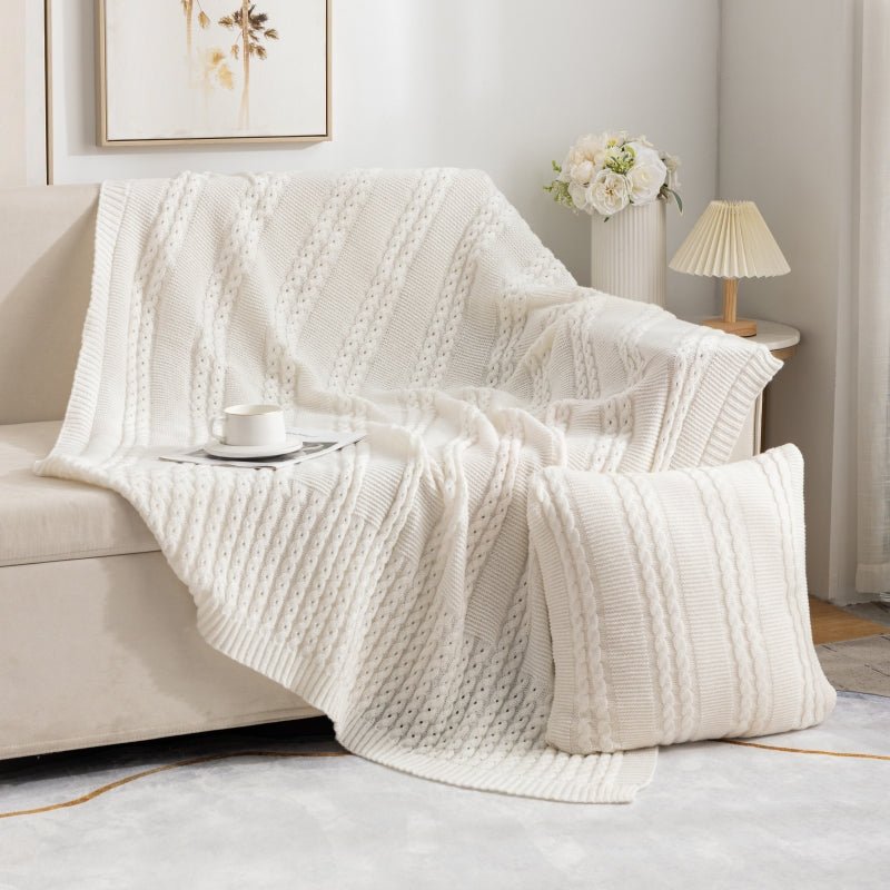 A beige Nordic knit throw blanket draped over the back of a sofa, with a cup of coffee on a tray and a lamp in the background.