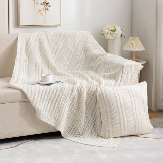 A beige Nordic knit throw blanket draped over the back of a sofa, with a cup of coffee on a tray and a lamp in the background.